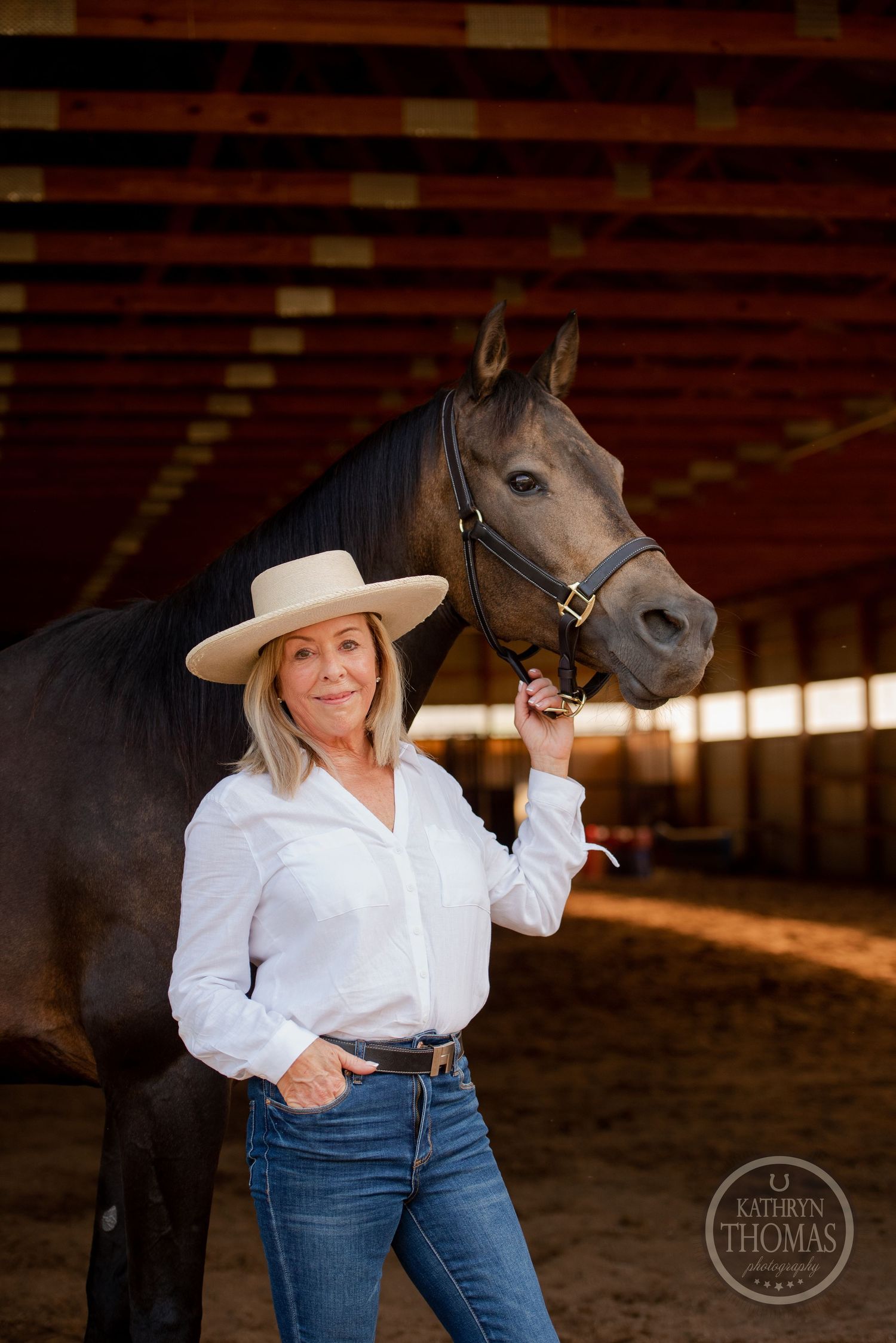 Equestrian in white shirt and wide-brimmed hat poses confidently with dark horse in arena.