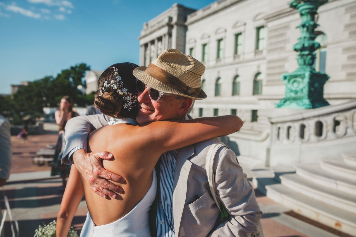 Guest in straw hat embraces someone during outdoor wedding celebration near historic building.