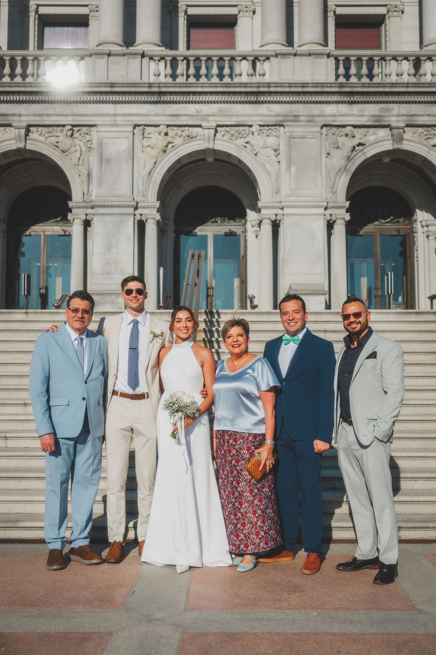 Wedding group poses in formal attire on steps of neoclassical government building.