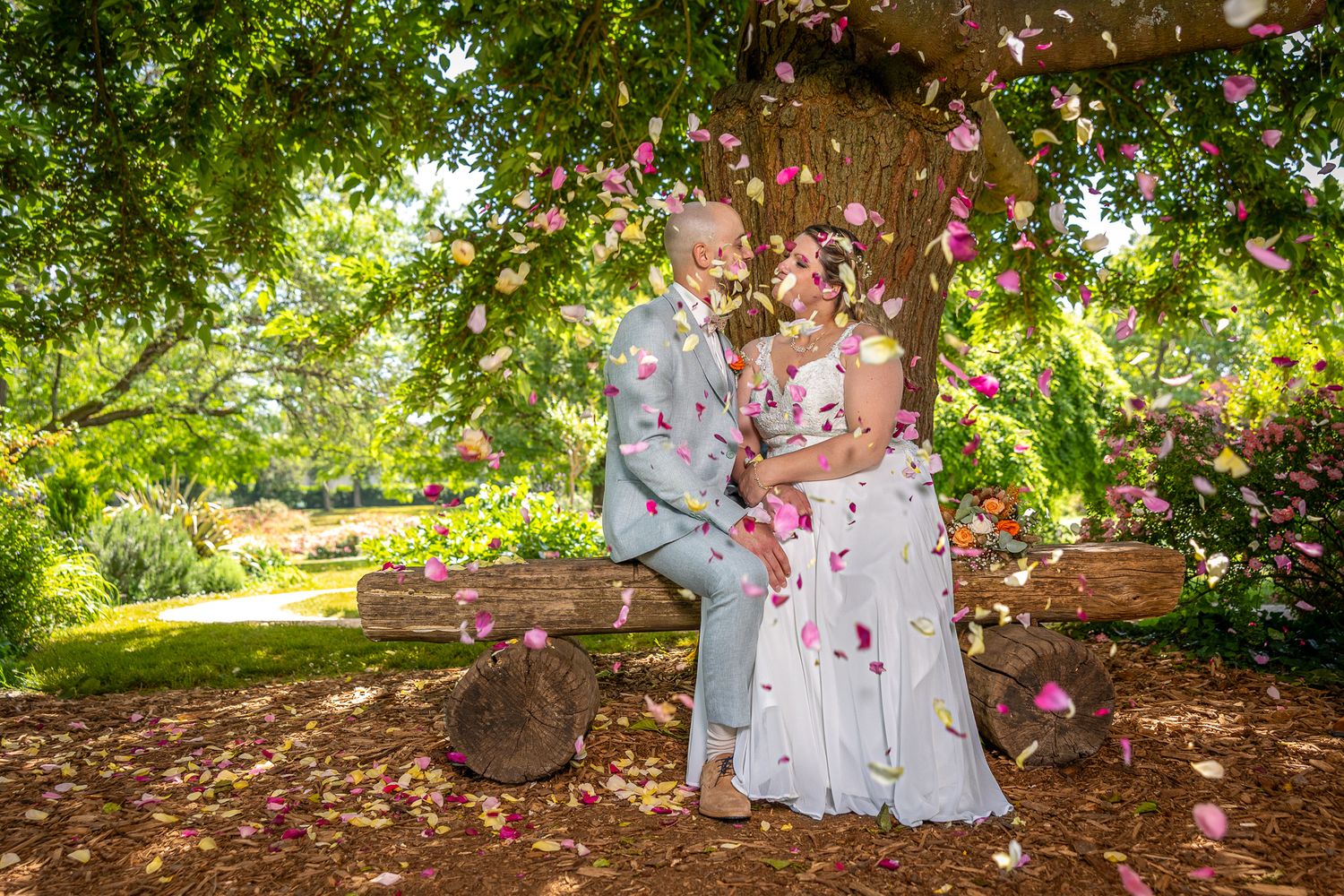 A couple stands under a tree while pink flower petals fall around them in a garden setting.