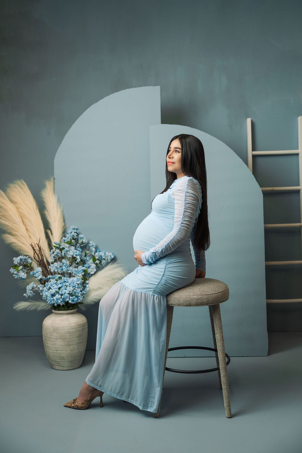 Pregnant woman in light blue lace dress sitting on stool beside floral arrangement in studio maternity photo.
