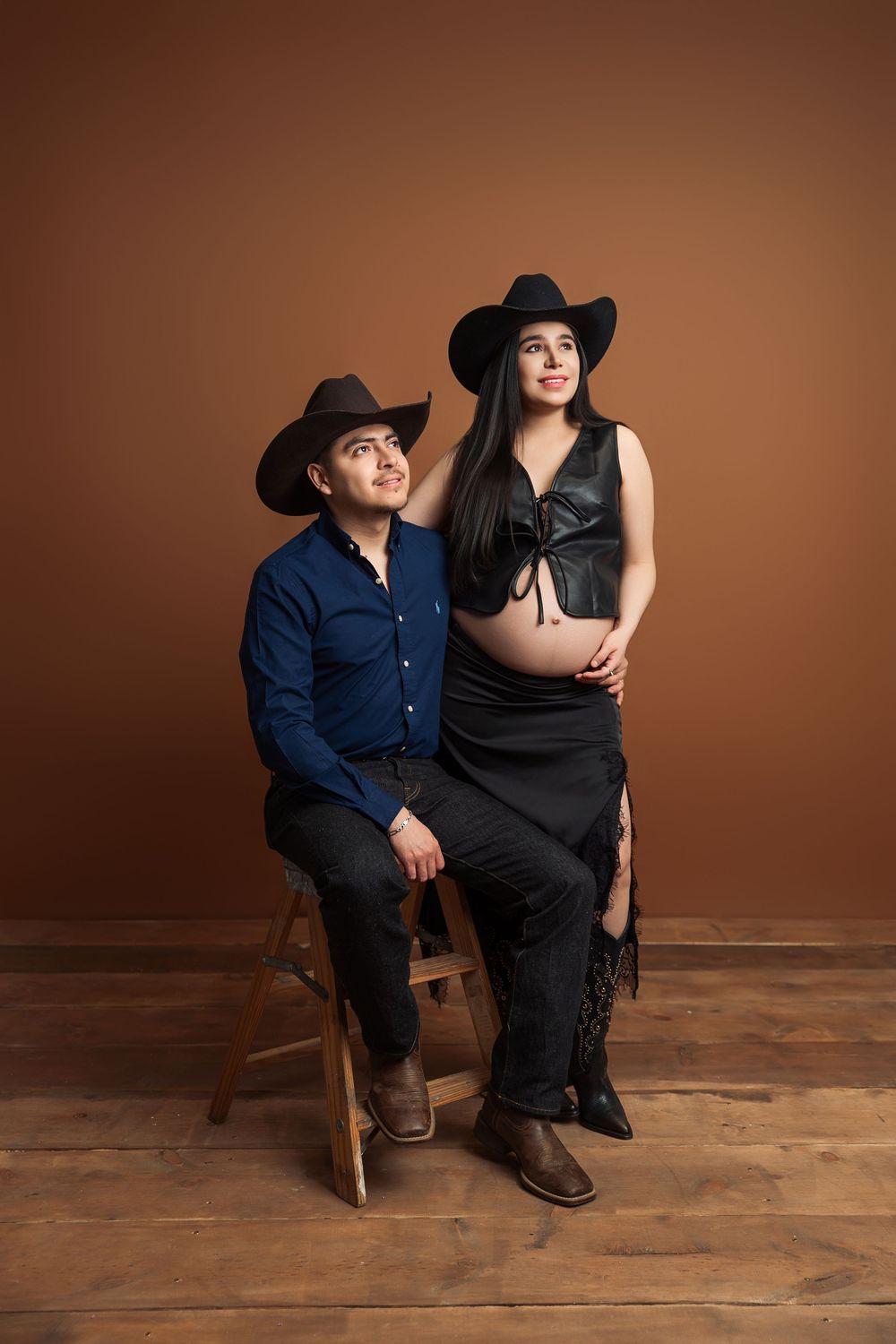 Couple in cowboy hats posing for maternity photo, man seated beside pregnant woman in black outfit, brown studio backdrop.