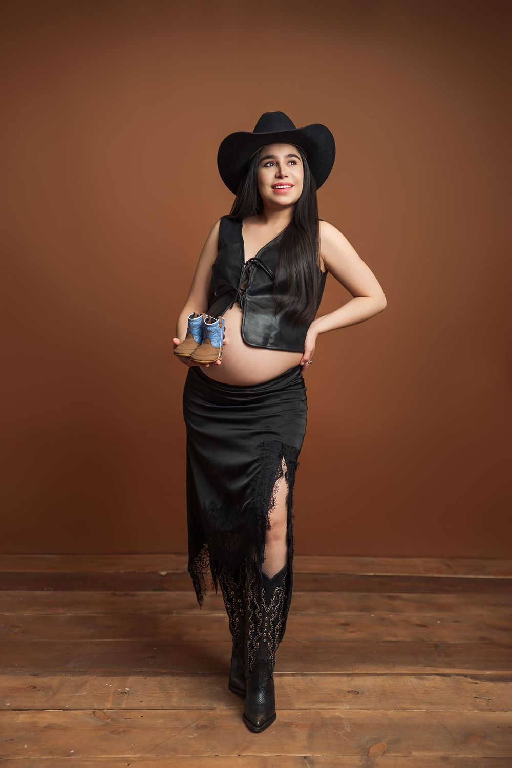 Pregnant woman in black cowboy hat, fringe skirt, and boots holding baby shoes against brown background.