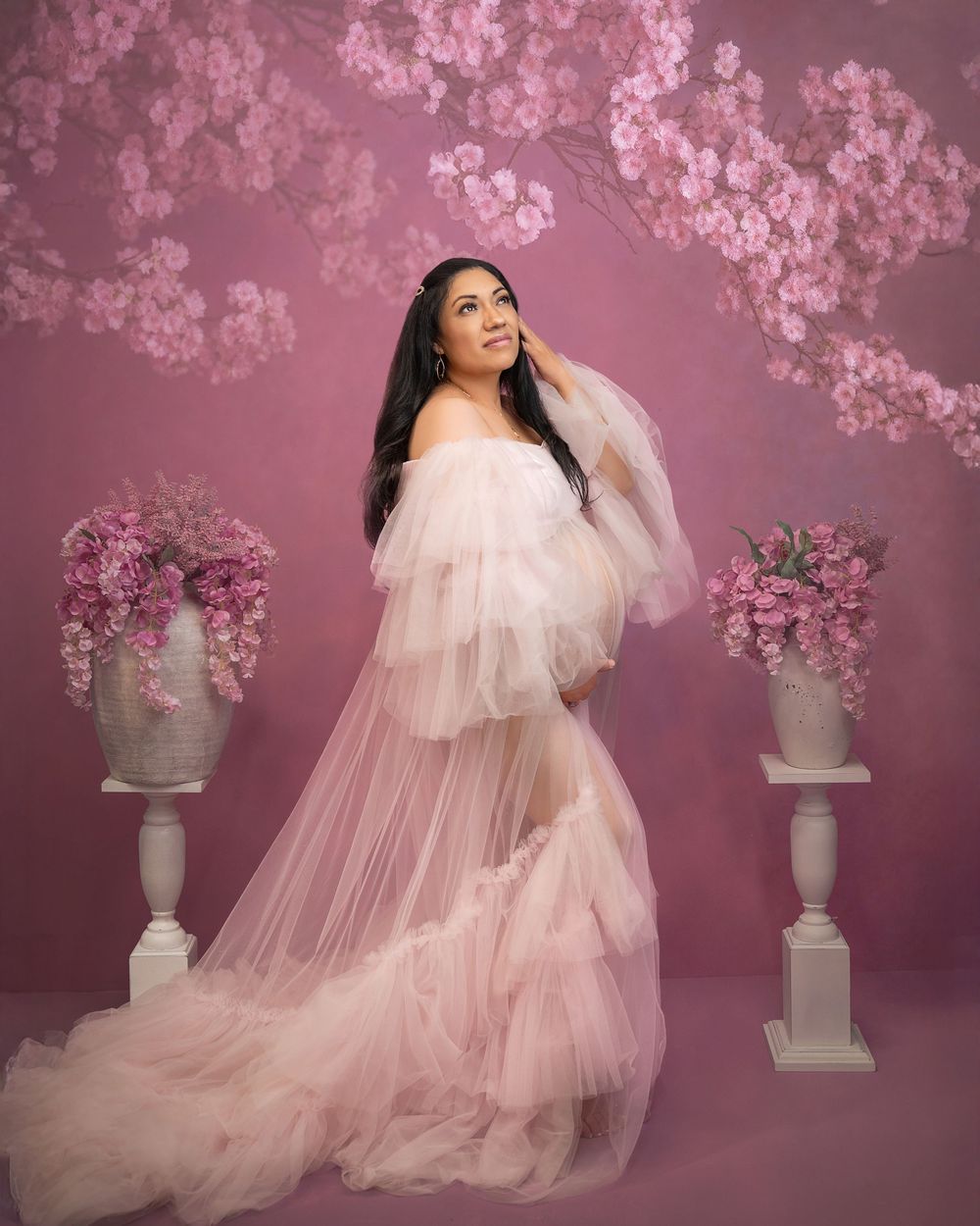 Pregnant woman in pink tulle gown poses gracefully before floral backdrop with cherry blossoms and flower arrangements.