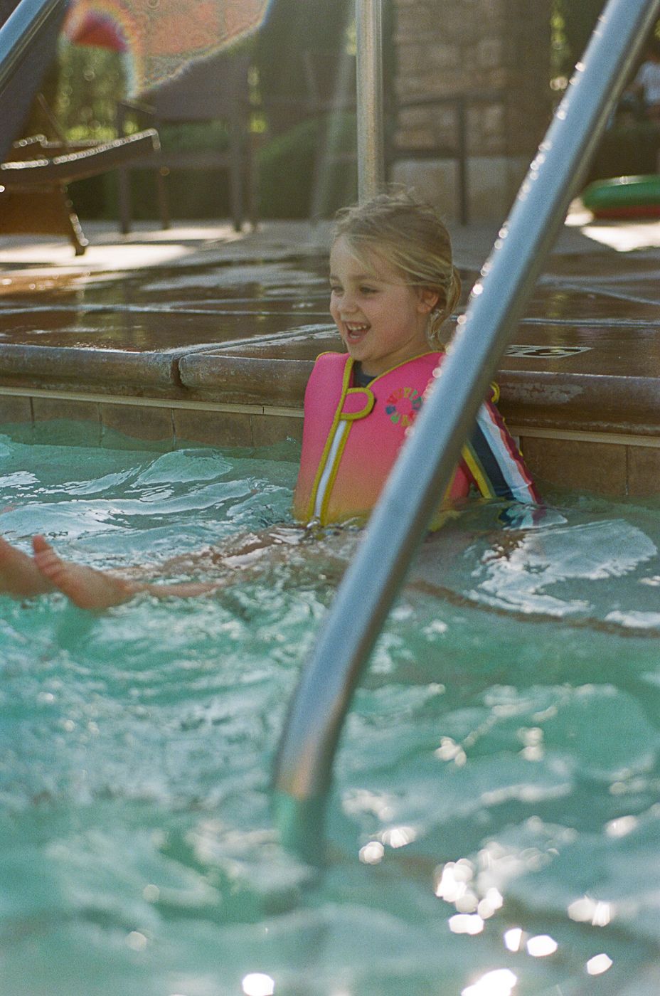 Child swimming and splashing in blue pool water with railing visible.