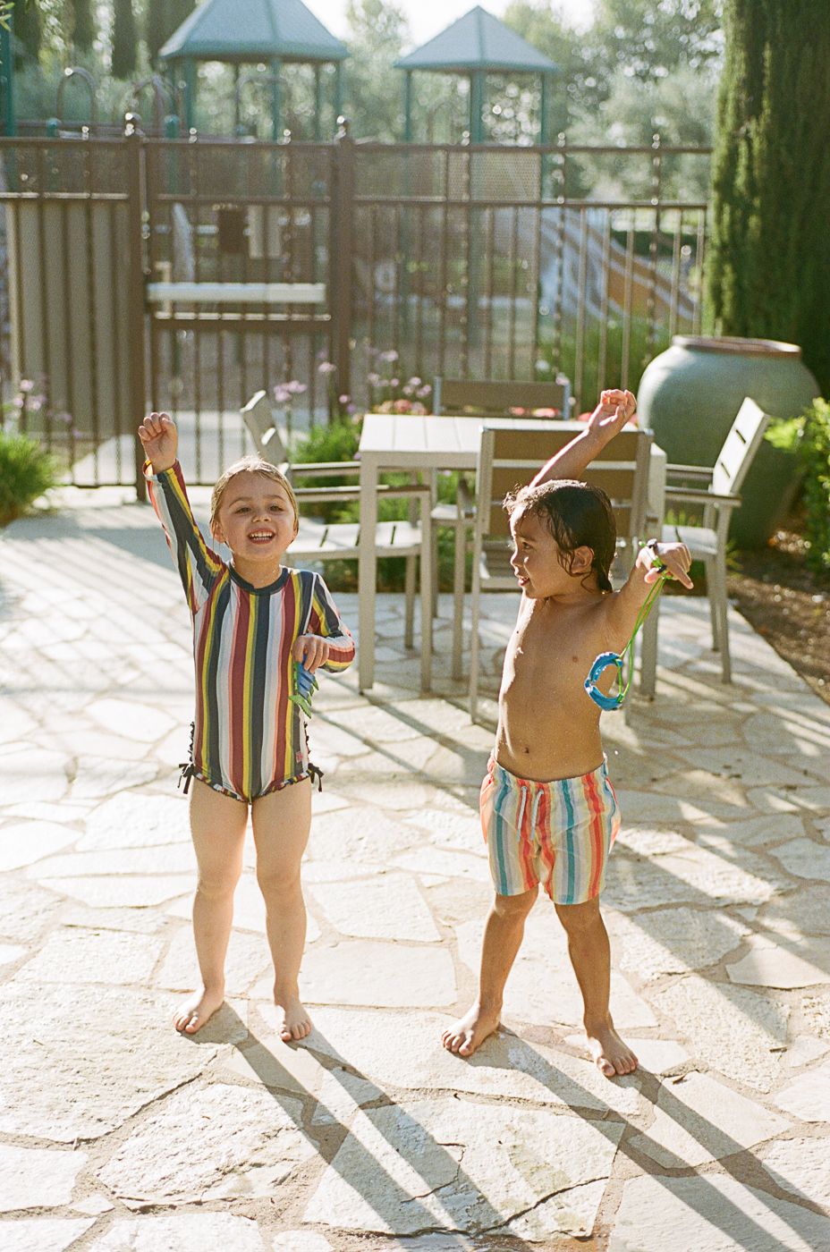 Children in colorful striped swimsuits playing in dappled sunlight on a patio.