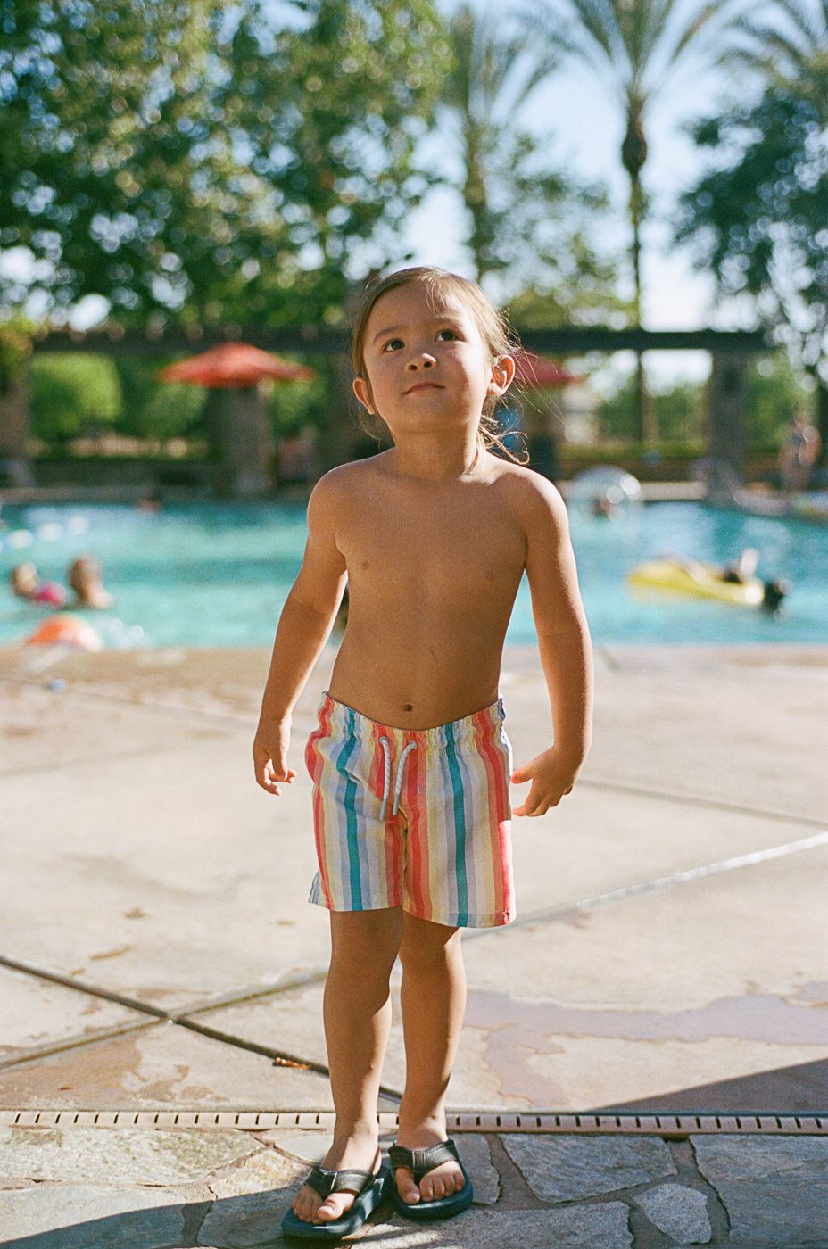 Young swimmer in striped swim shorts stands by pool edge on sunny day.