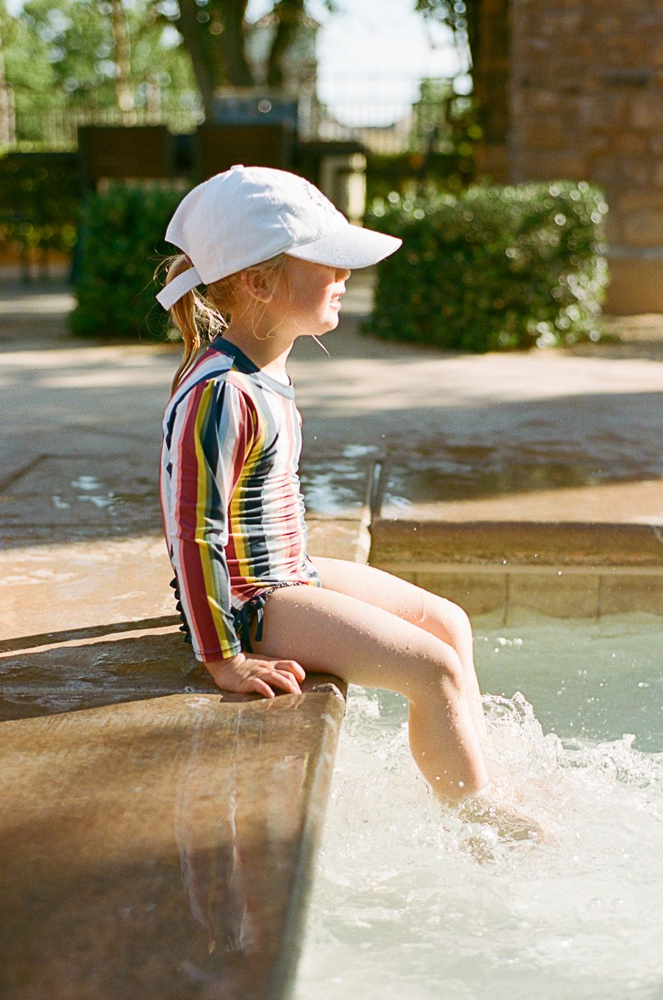 Young children in striped swimsuits dangling feet in sparkling pool water on a sunny summer day.