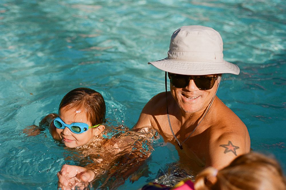 People swimming in crystal clear blue pool water wearing sunhats and goggles.