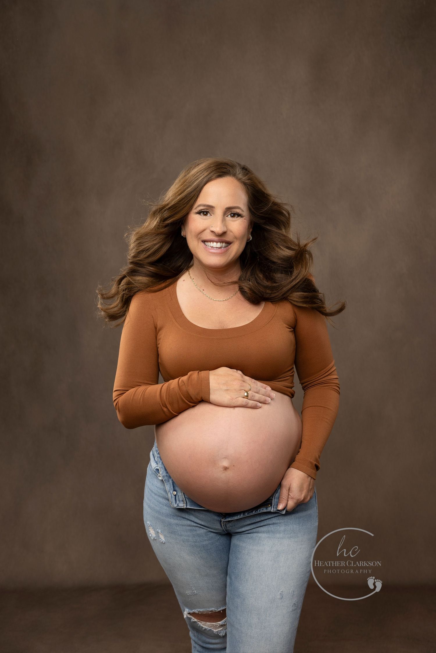 Maternity portrait of woman in brown top and jeans against dark backdrop.