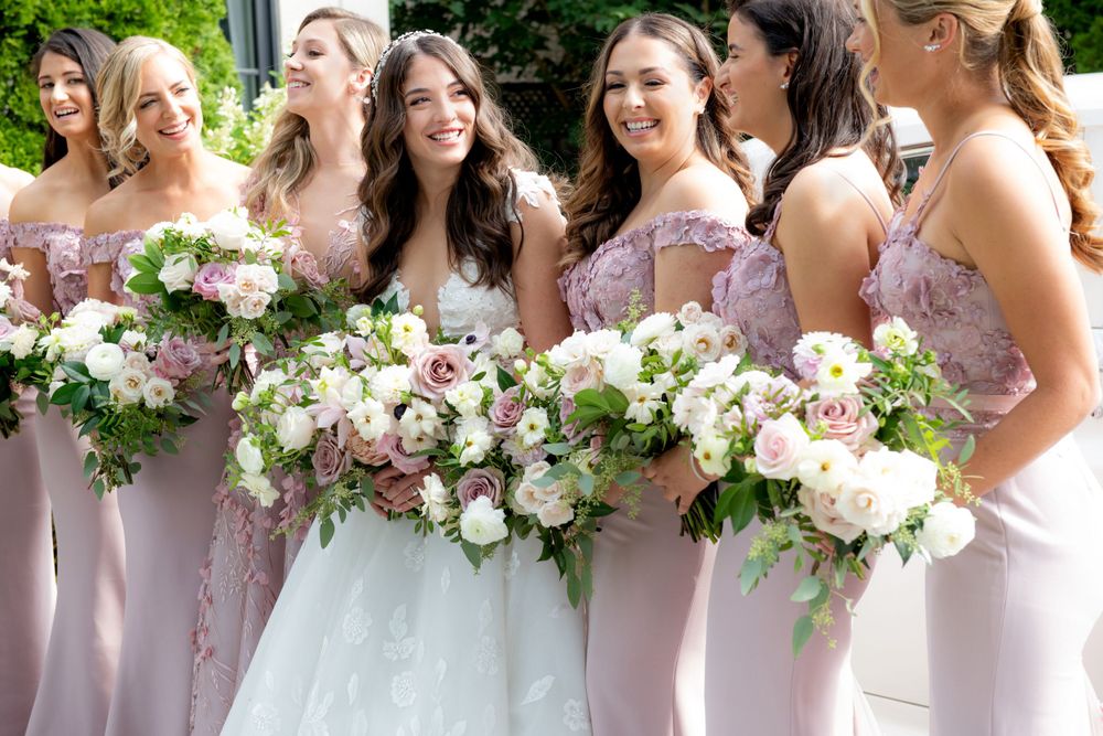 Bridal party in soft pink dresses holding lush floral bouquets of roses and greenery.