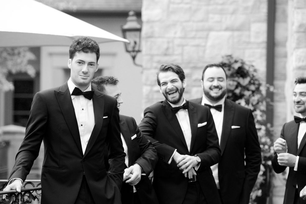 Group of groomsmen in black tuxedos sharing a laugh outdoors.