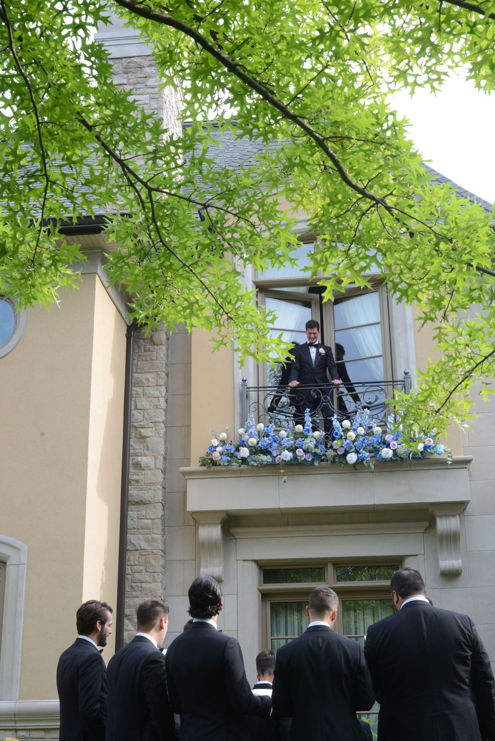 Groomsmen looking up at someone standing on a balcony decorated with blue flowers.