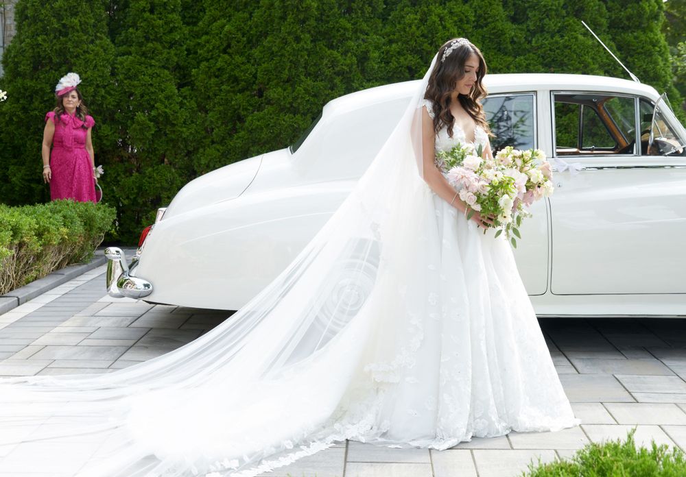 A bride in a flowing white gown stands beside a classic white car on a sunny day.