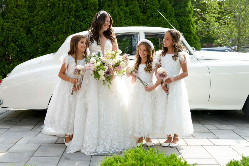 A classic white wedding party poses by a vintage car with white flower bouquets.