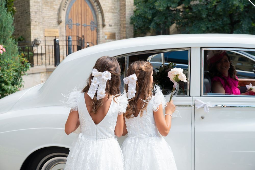 Three attendants in white dresses peek into a vintage car window.