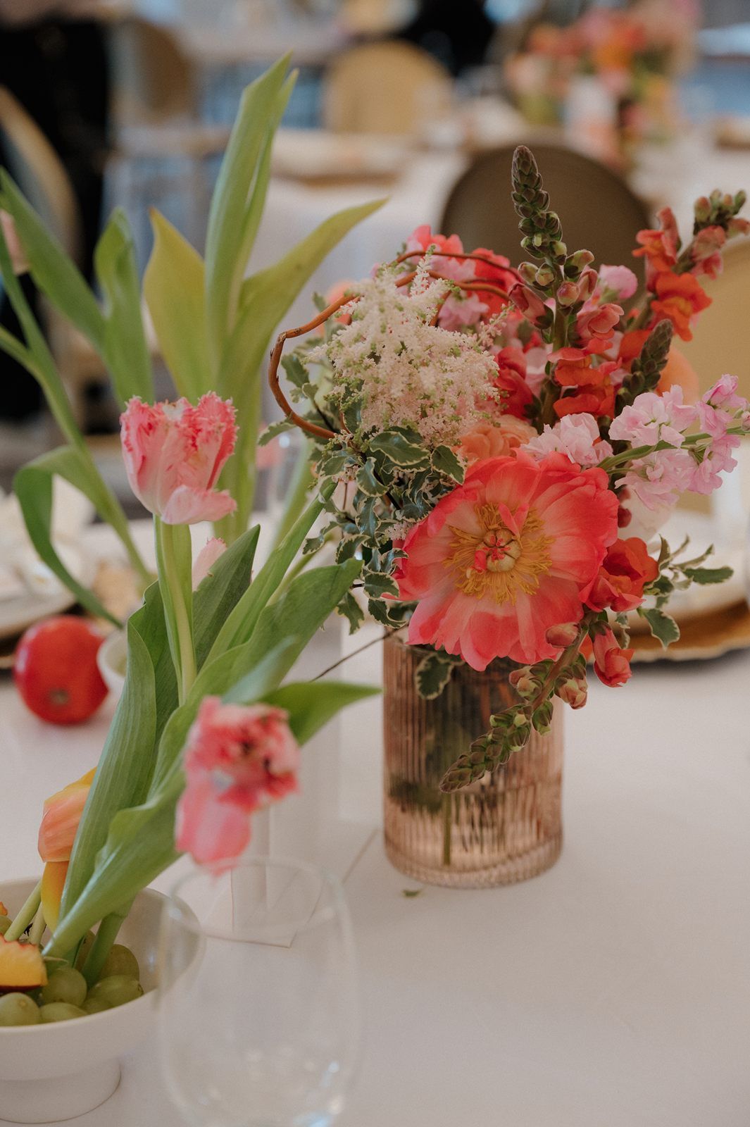 A romantic table setting with pink tulips and coral flowers in glass vases against a soft blurred background.