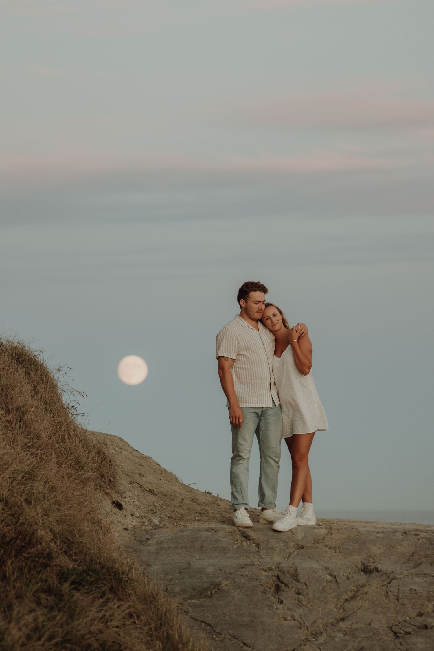 Couple embracing on sandy coastal dunes with glowing full moon in soft twilight sky.