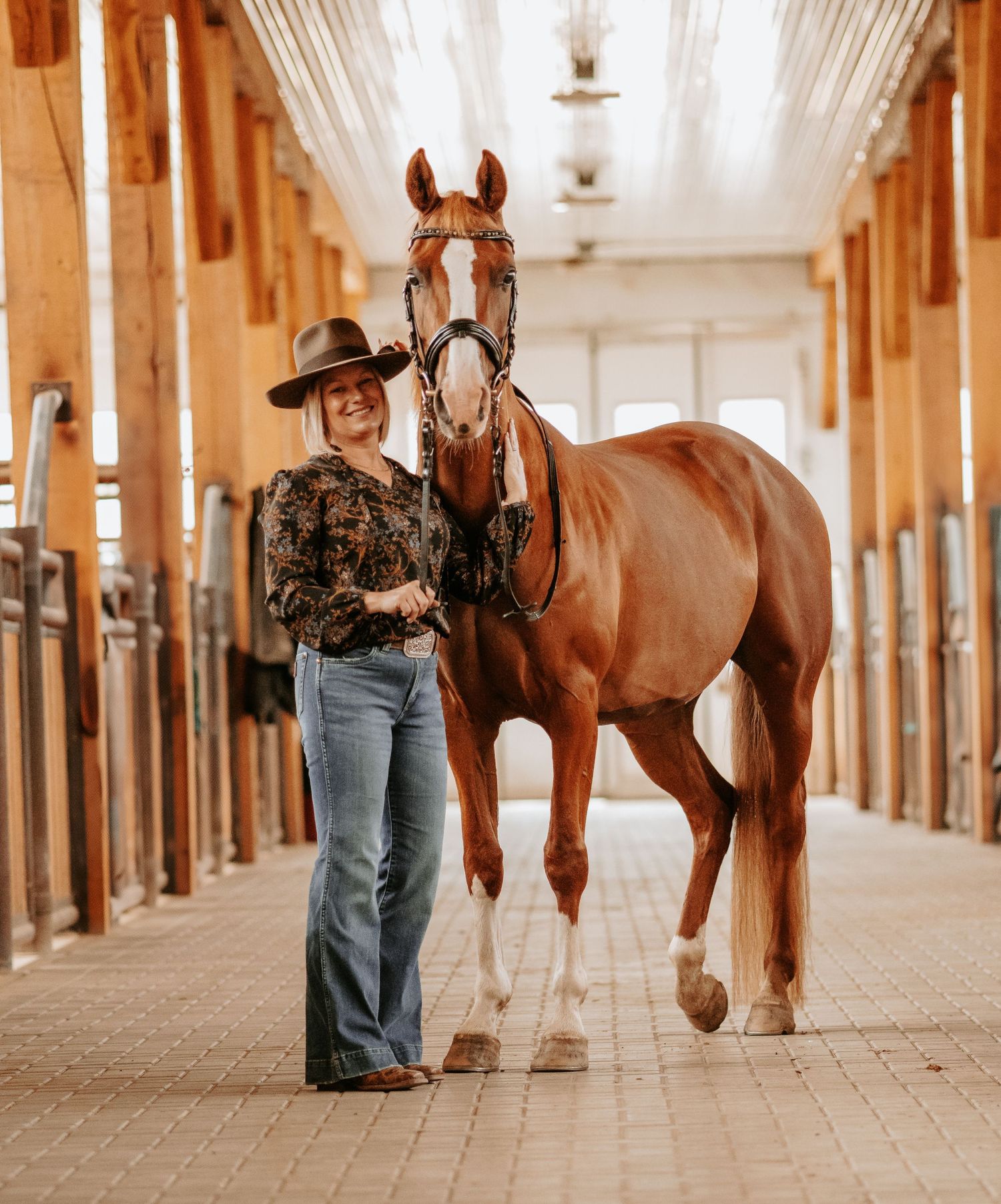 Woman in cowboy hat walks a chestnut horse through a sunlit wooden stable aisle.