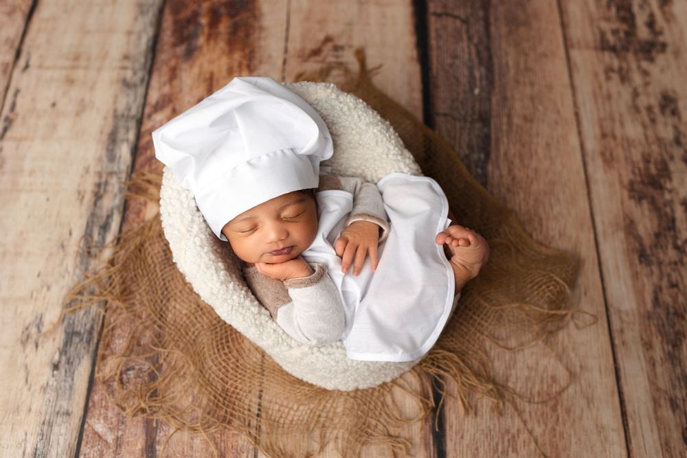 Sleeping newborn wearing white chef hat in cozy basket on wooden floor.