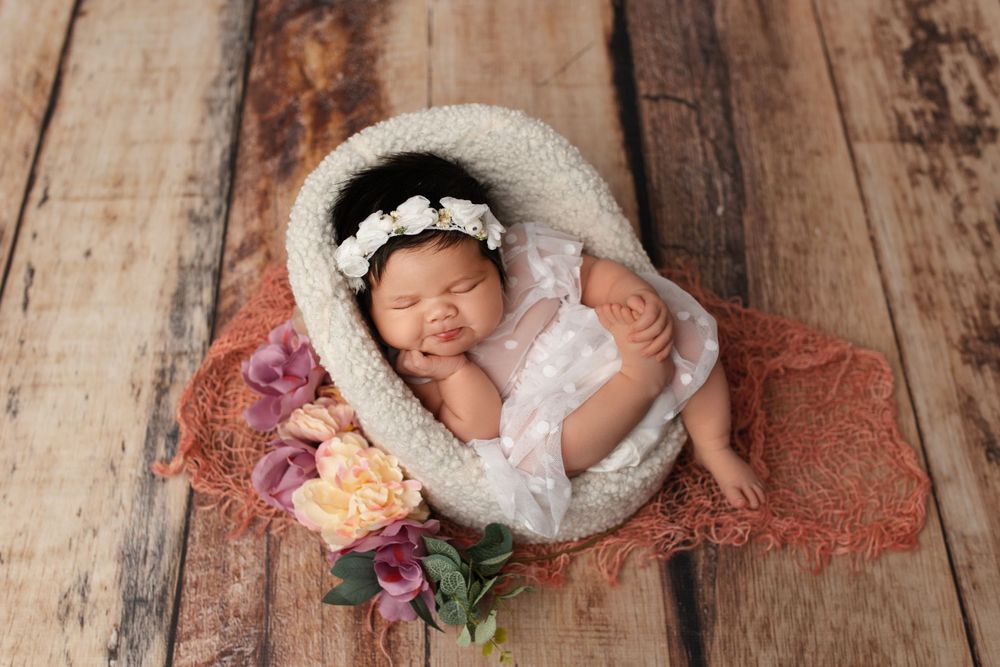 Newborn baby sleeping in white cozy nest with floral accents on wooden backdrop during professional photoshoot.