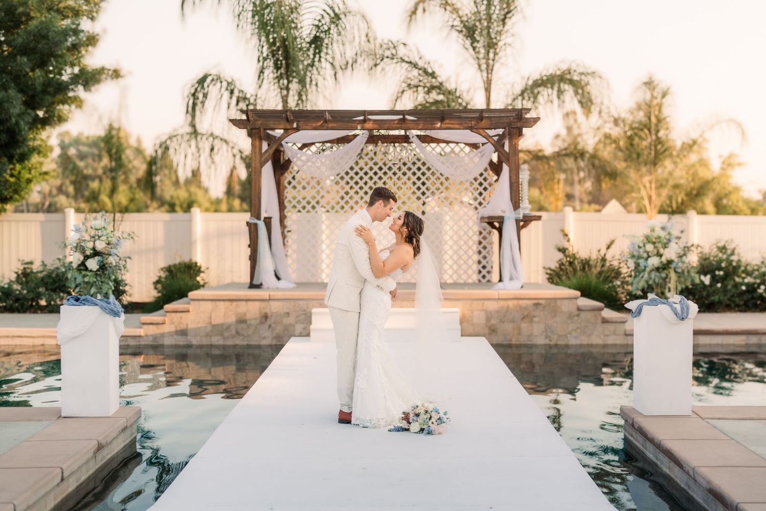 A bride poses elegantly near white pillars and palm trees at an outdoor venue.