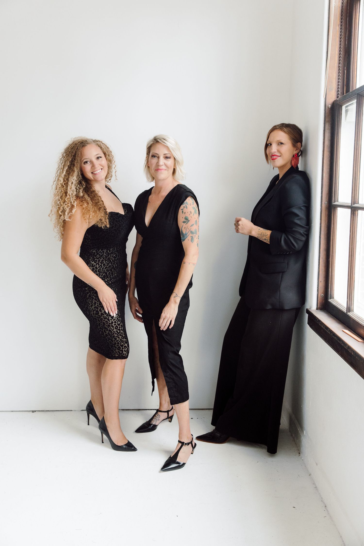 Three women in elegant black attire posing together against a white wall.