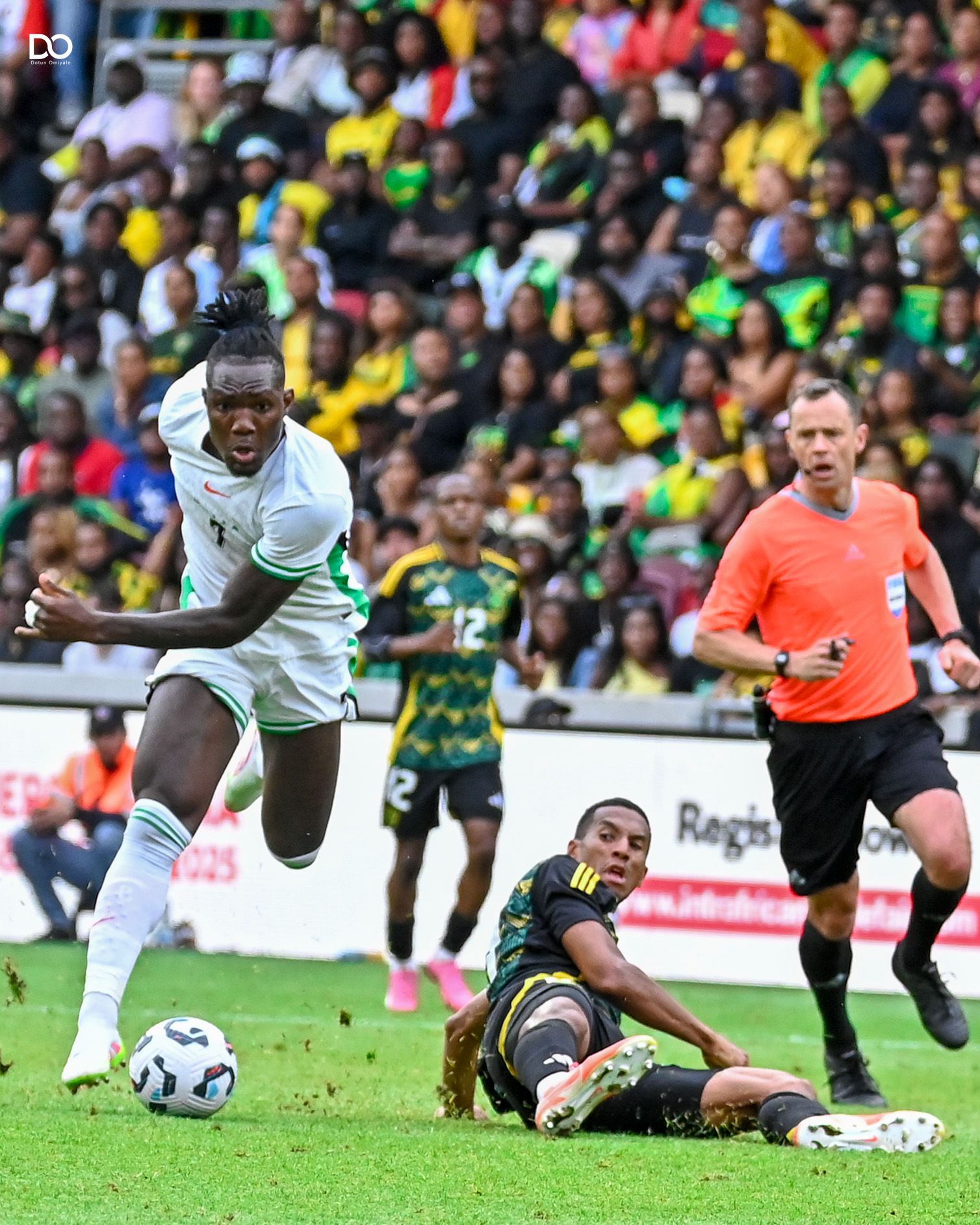 Nigerian Soccer player in white jersey leaps over fallen opponent during match with colorful crowd in background.