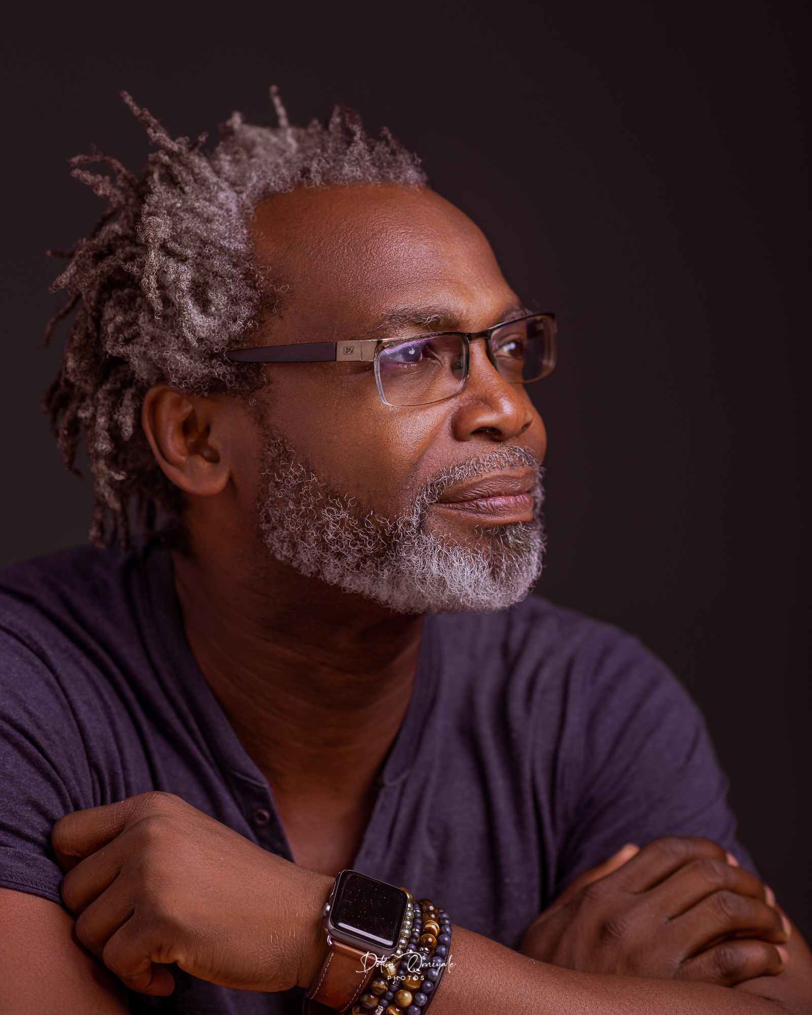 Portrait of a distinguished person wearing glasses and a purple shirt, with silver hair styled in dreadlocks and a salt-and-pepper beard.