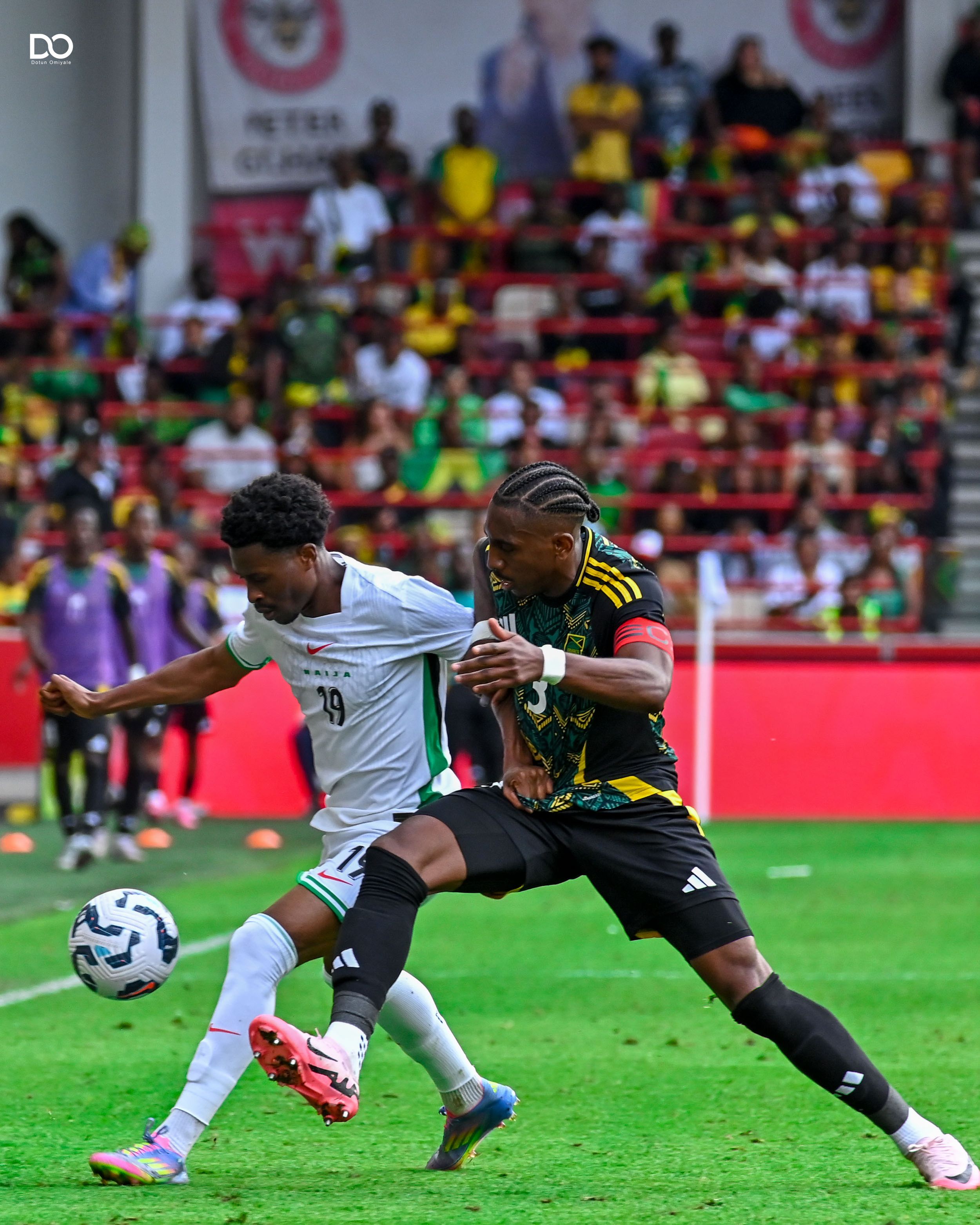 Two soccer players battle for possession of the ball during an intense match with colorful crowd in background.