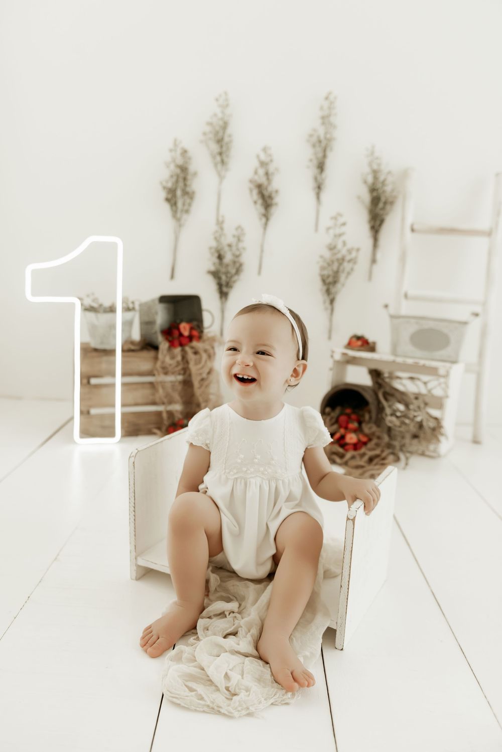 Baby in white dress sits and smiles on light wooden floor with pampas grass decor in background.