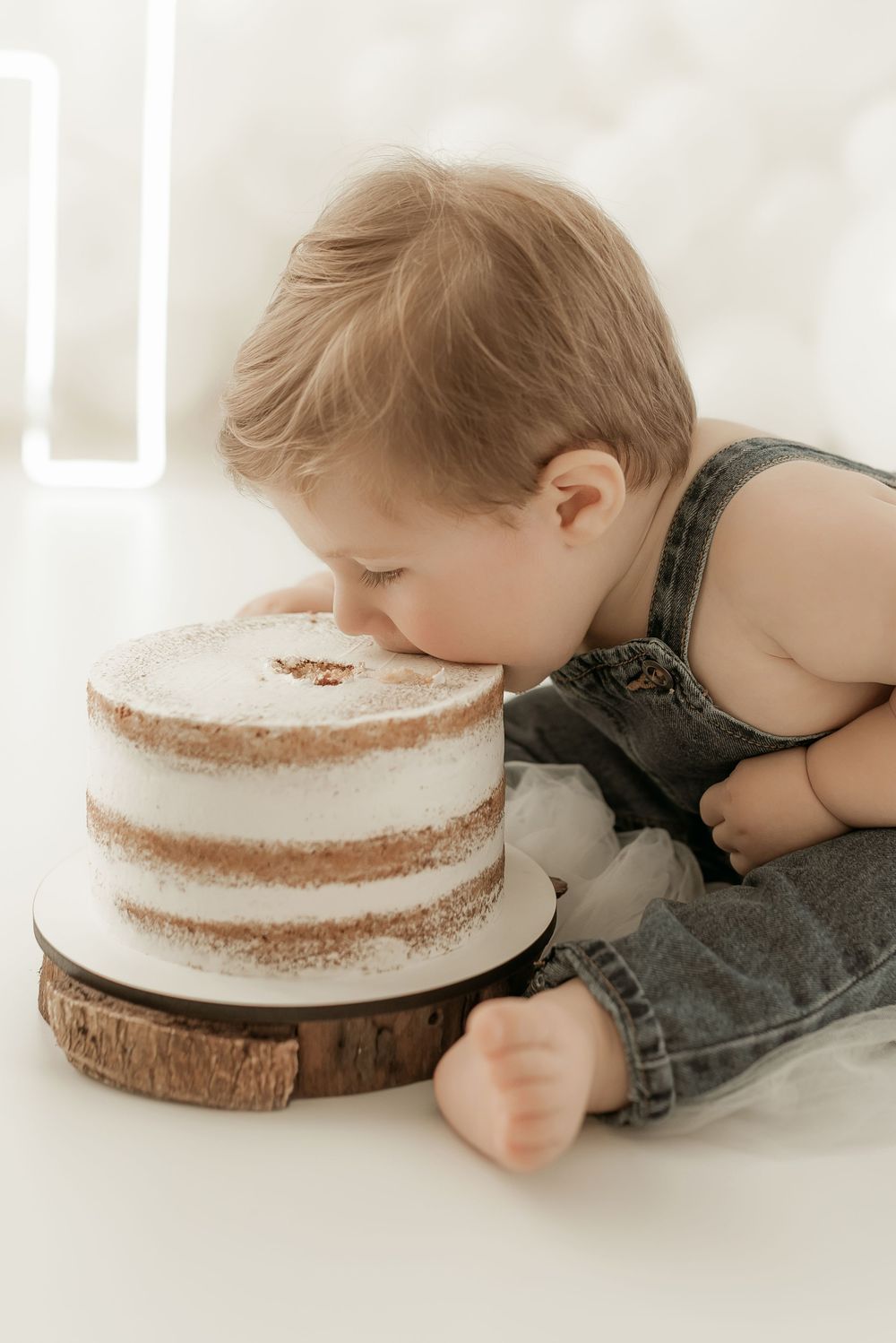 Baby in denim overalls eagerly takes a bite of a naked layer cake displayed on a rustic wood slice.
