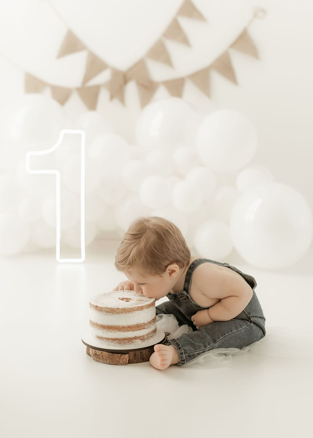 Small child in suspenders leans down to inspect a naked white birthday cake with number 1 decoration on top.