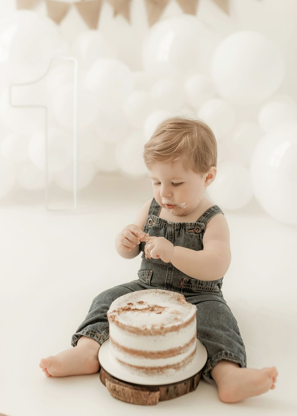 A small child in denim overalls sits barefoot next to a rustic white naked cake with bunting decorations in the background.