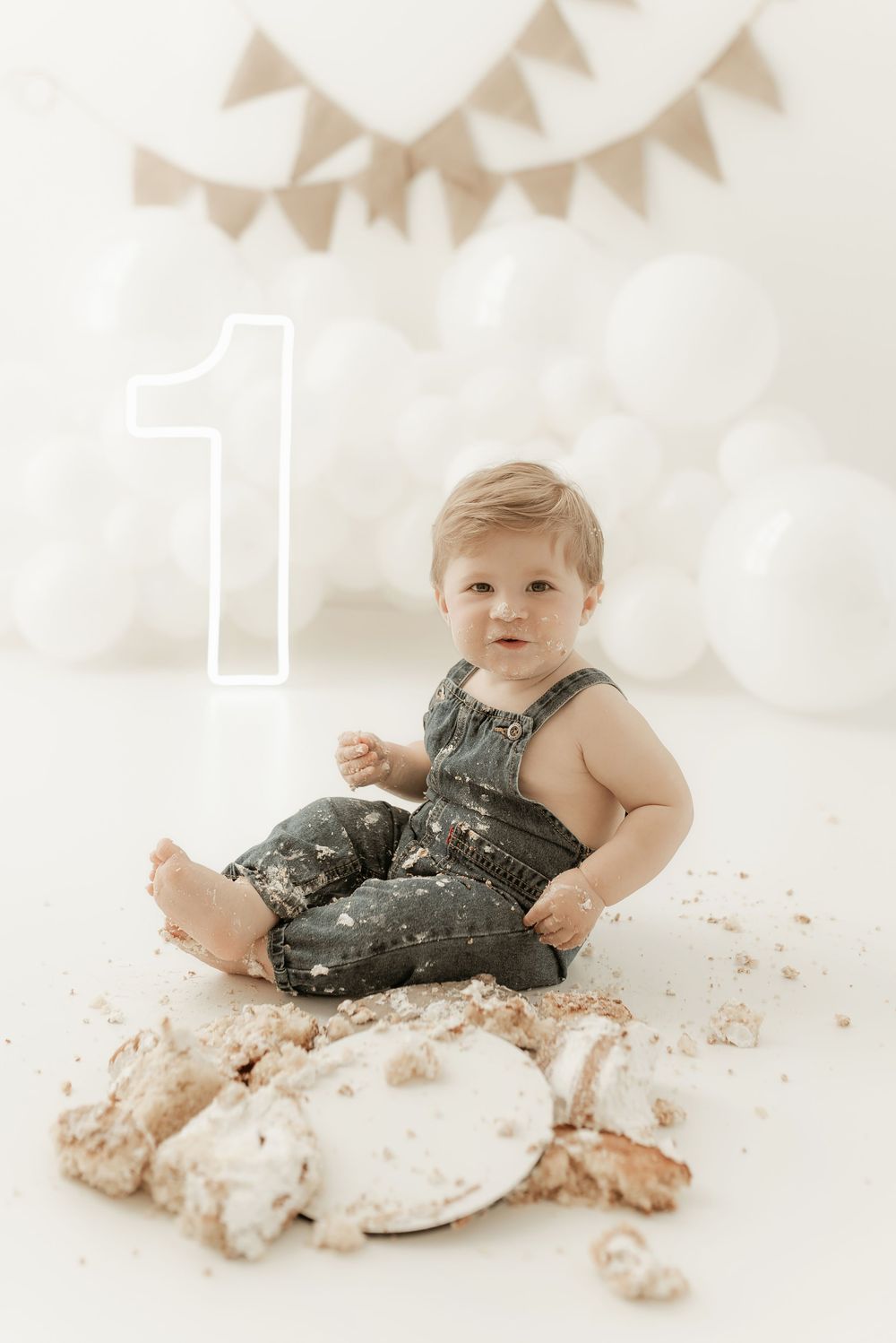 First birthday cake smash photo session with a baby in green overalls sitting among smashed white cake on light background.