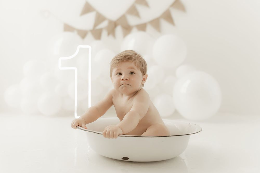 A baby sits in a white ceramic bowl against a soft white background with delicate bunting decorations.