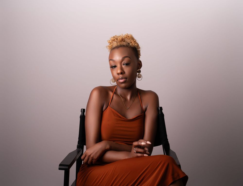 A portrait of a person in an orange dress seated in a directors chair against a neutral backdrop.