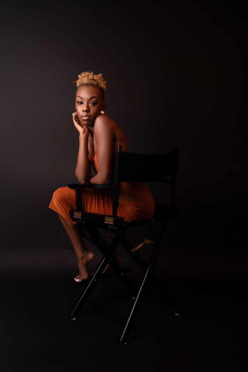 An artistic portrait of a person in orange attire posed on a directors chair in dramatic lighting.