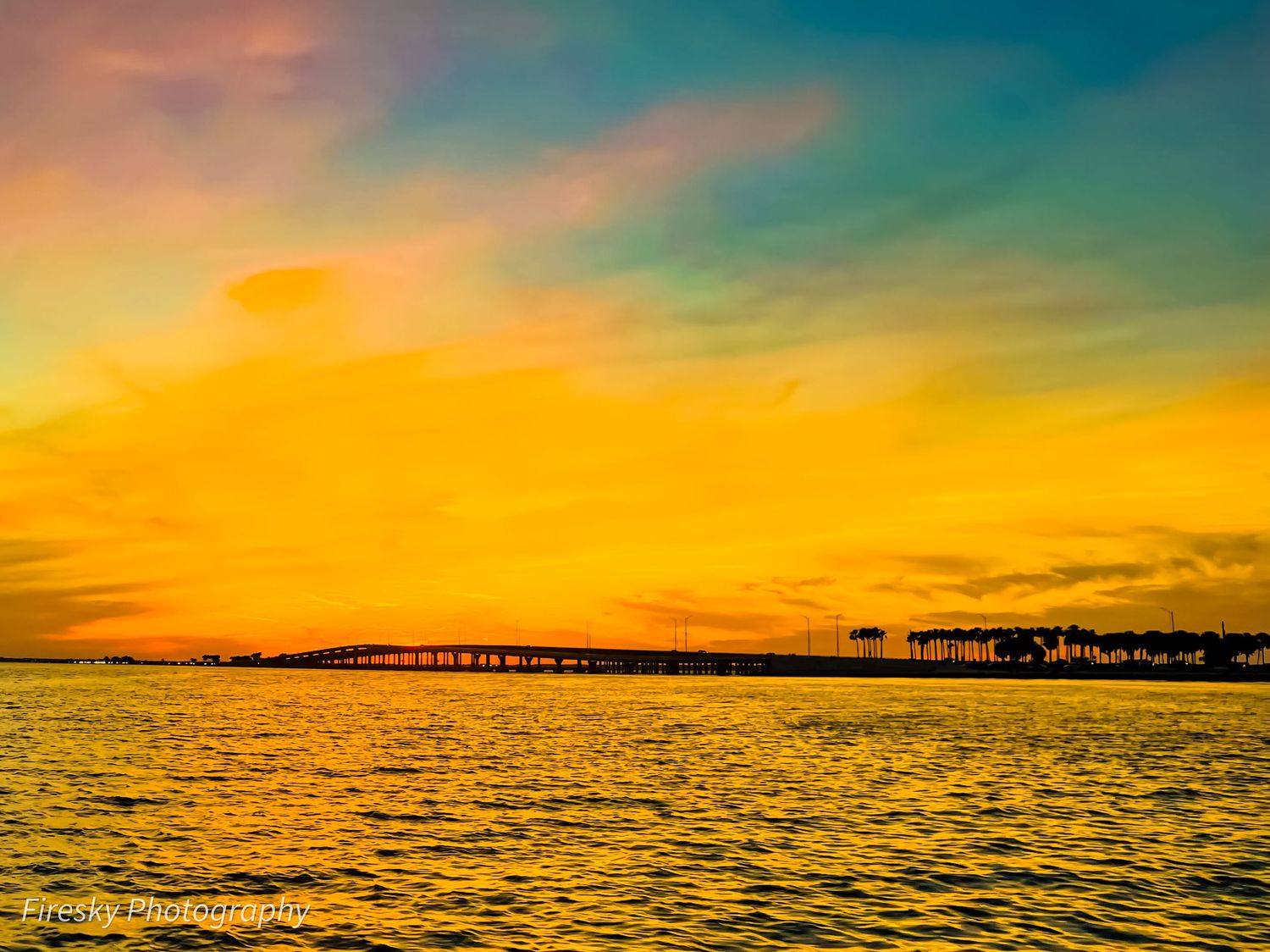 Vibrant sunset over water with silhouetted bridge and palm trees in the distance.