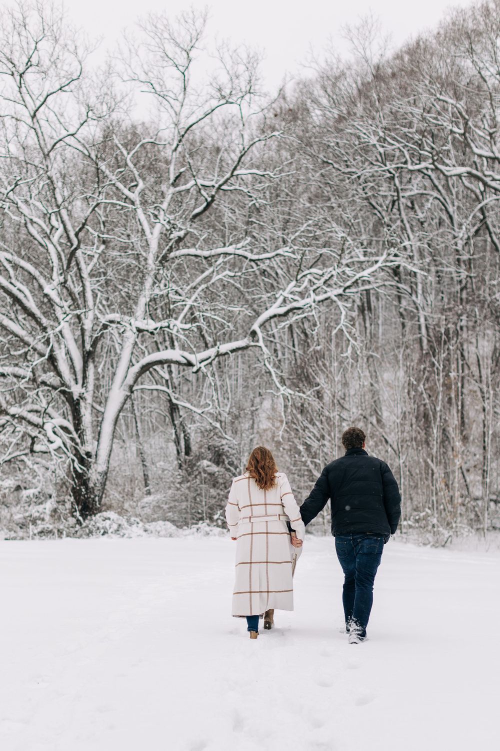 Couple walks hand in hand through snowy winter forest landscape.