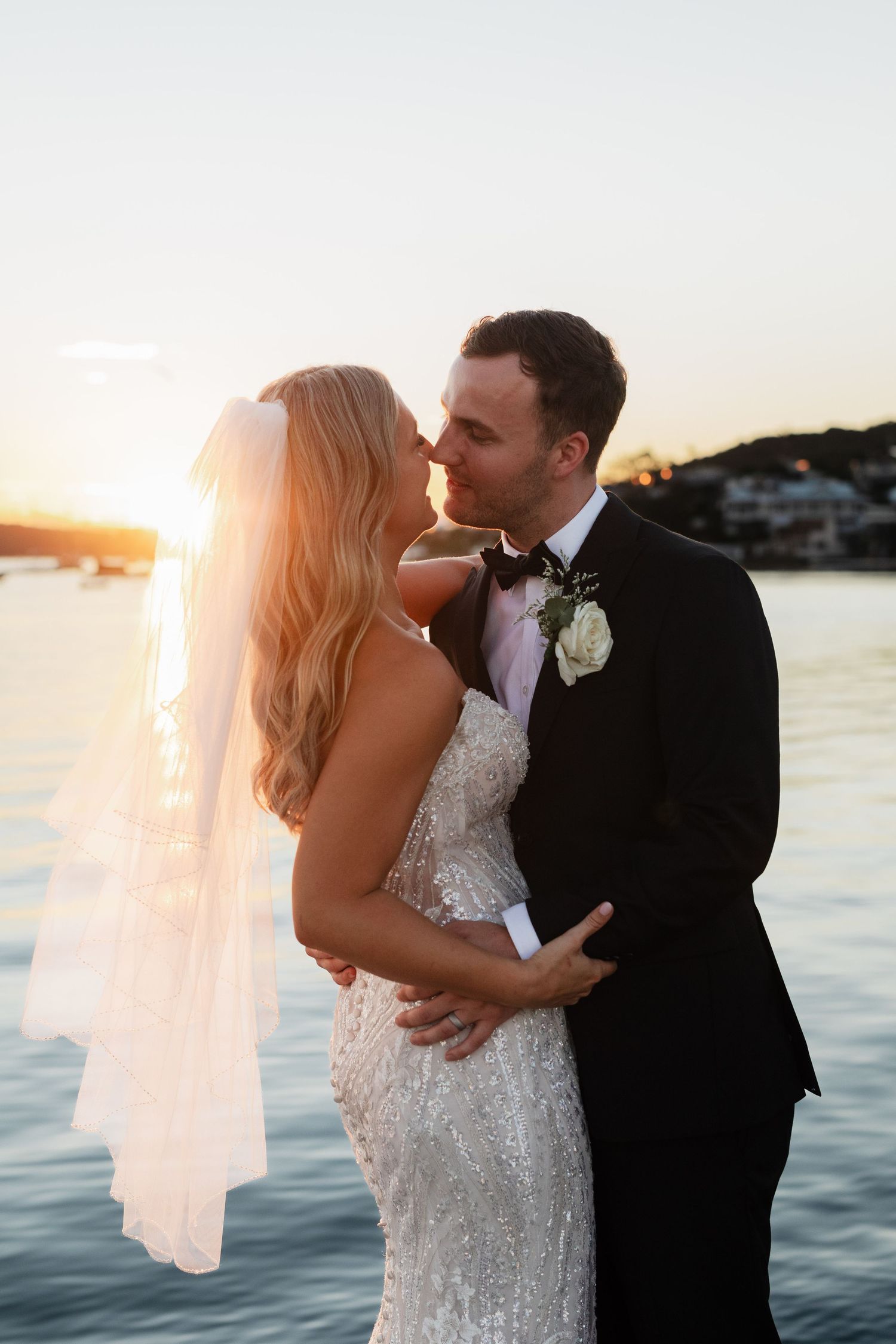 Sunset wedding portraits by the water with bride's veil flowing in the breeze.