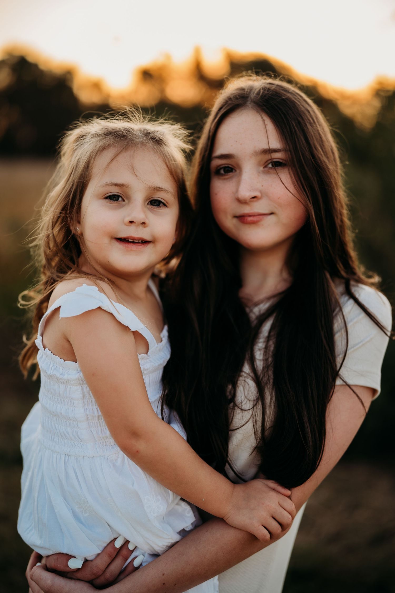 Older sister with long dark hair hugging younger blonde sister at golden sunset.