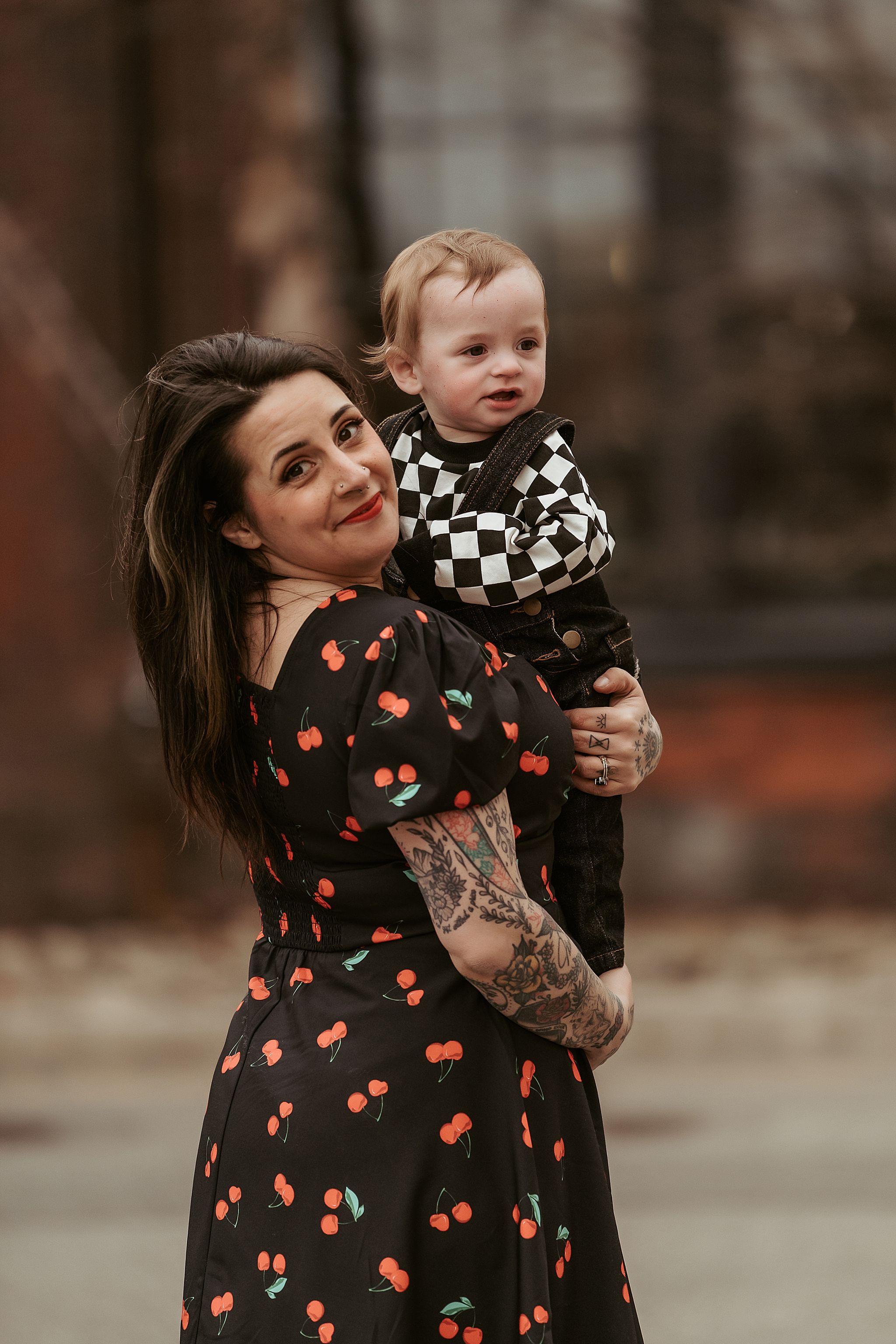 Mother in cherry print dress holds toddler in checkered outfit against autumn backdrop.