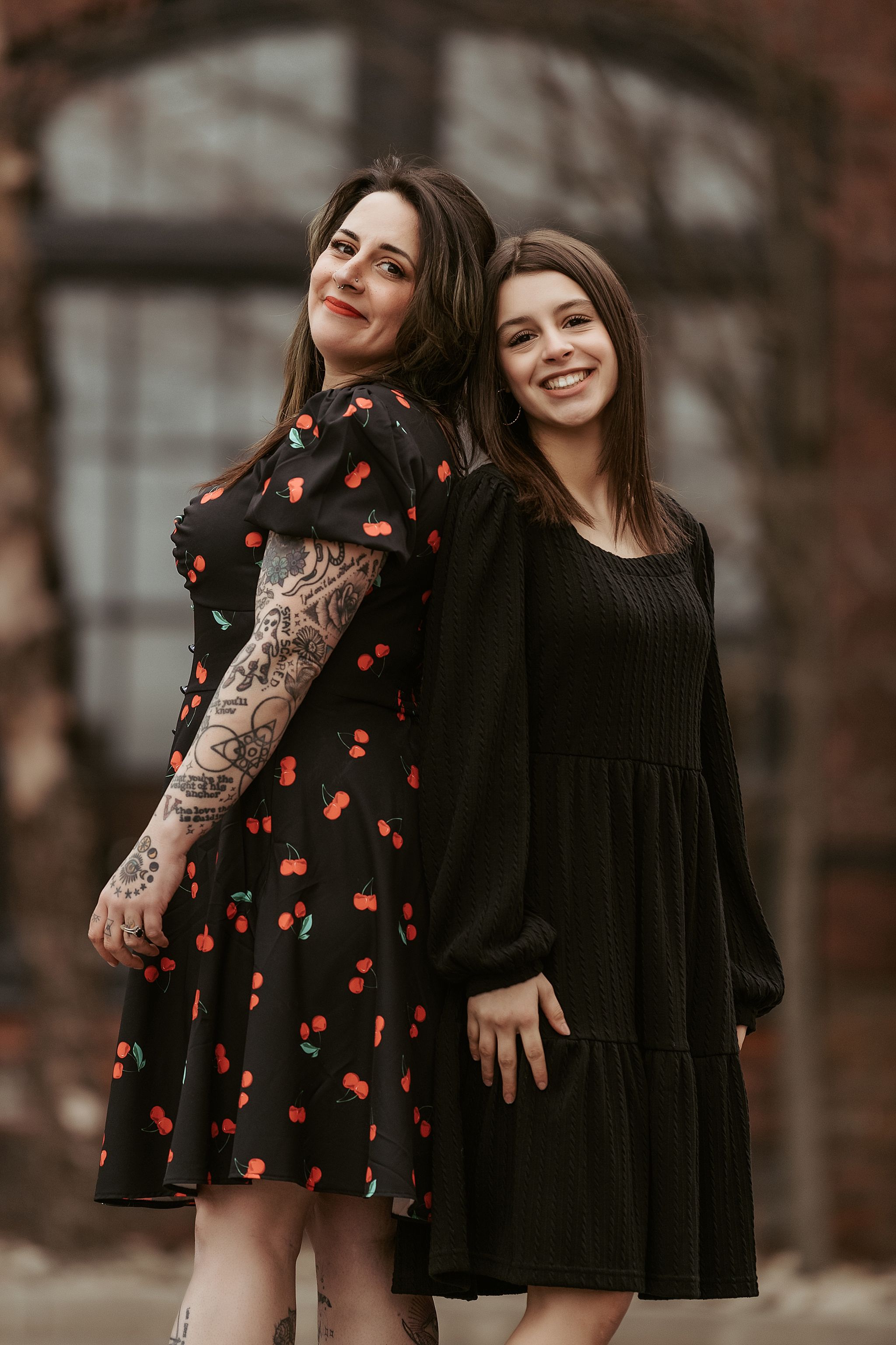 Two women pose together against brick building, one wearing black dress and one in cherry print dress.