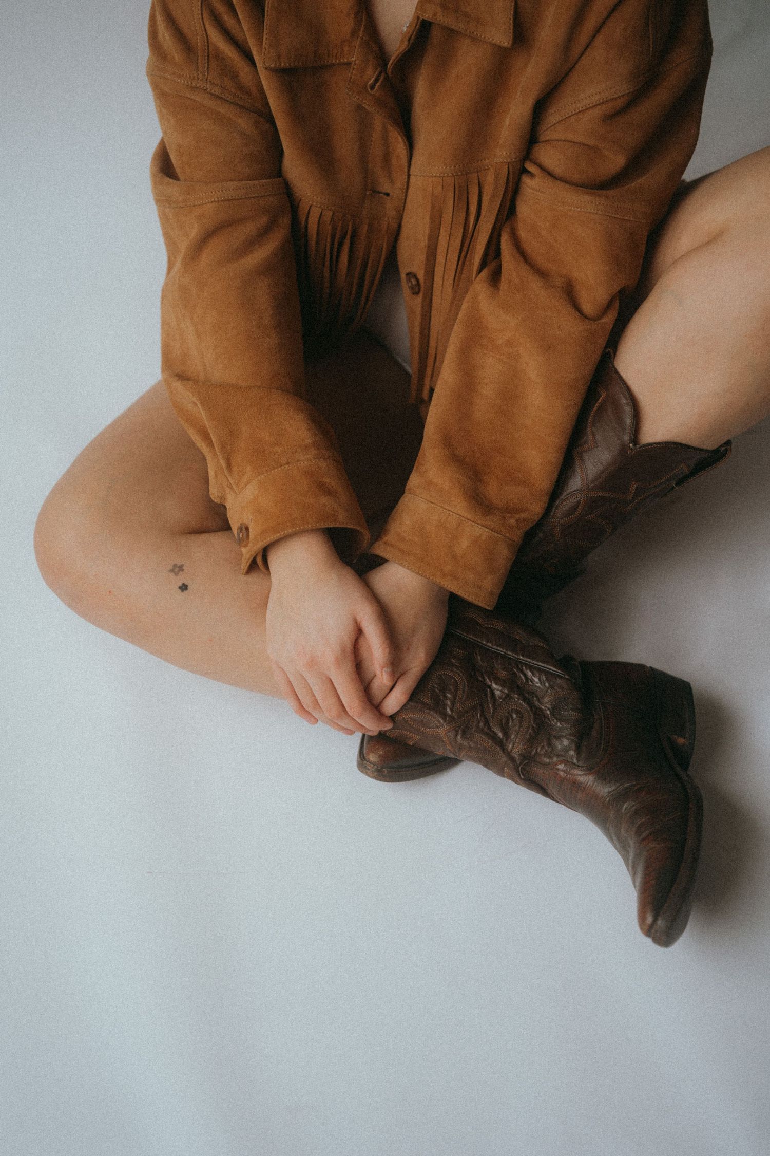 Person wearing brown shirt and cowboy boots sits cross-legged on white floor.