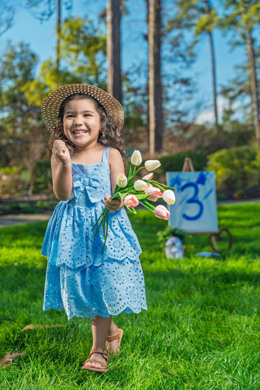 Happy little girl in blue dress and straw hat holding pink tulips outdoors near a number 3 birthday sign.