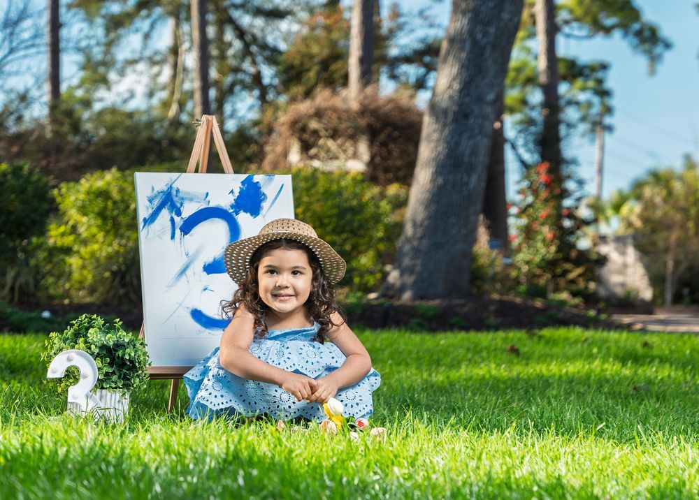 Toddler girl in blue dress and straw hat sits on grass beside a canvas painting the number 3 for her birthday.