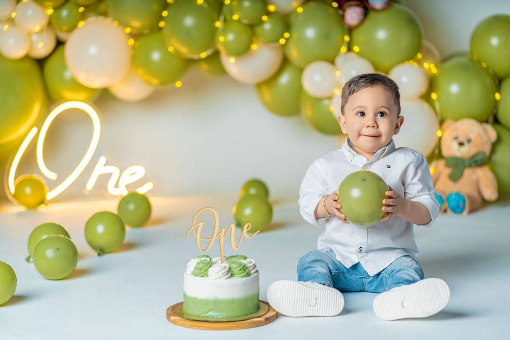 Baby boy holds green balloon at his first birthday party with green cake and balloon arch backdrop.