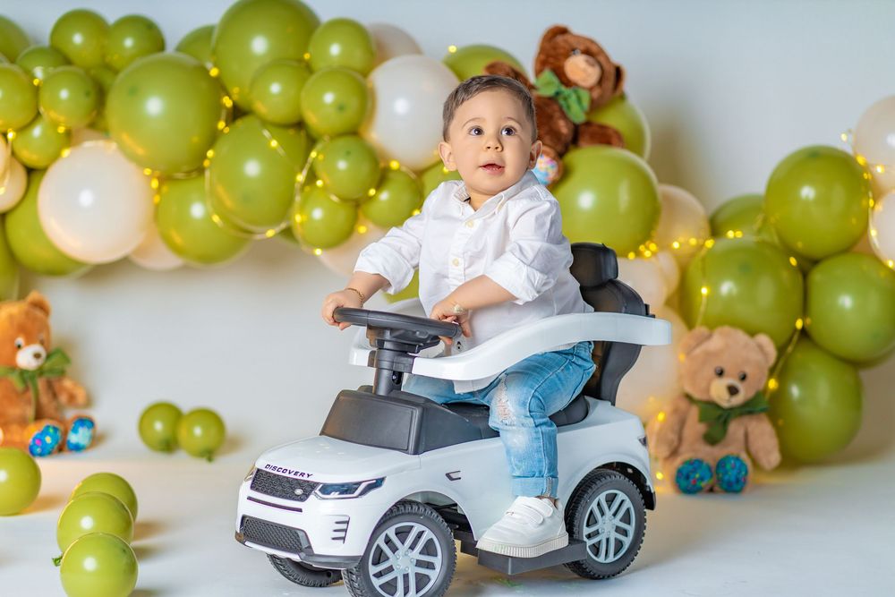Toddler boy in white shirt rides toy Range Rover car surrounded by green balloons and teddy bears at birthday party.