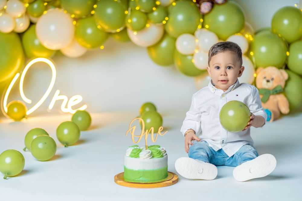 A baby boy celebrates his first birthday with a green cake and balloon arch backdrop.